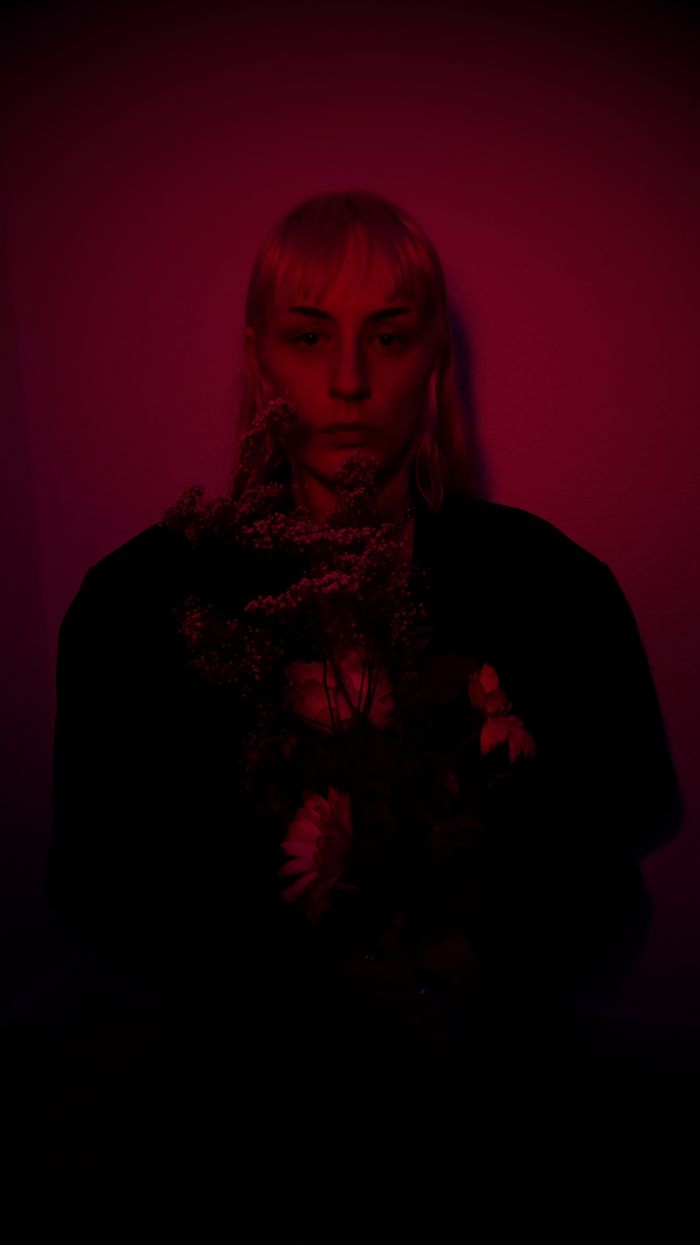 Serious woman with blond hair in dark clothes standing with sprig on plant in studio illuminated with dark red light and looking at camera
